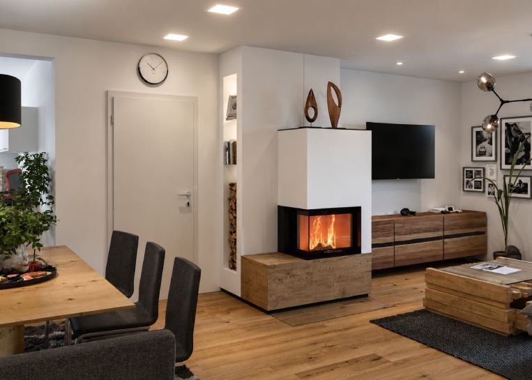 Gesamtansicht eines Neocube Modul-Ofens mit Ecksichtscheibe, eingebettet in ein weißes Wandregal mit Holzlager und Dekofächern.