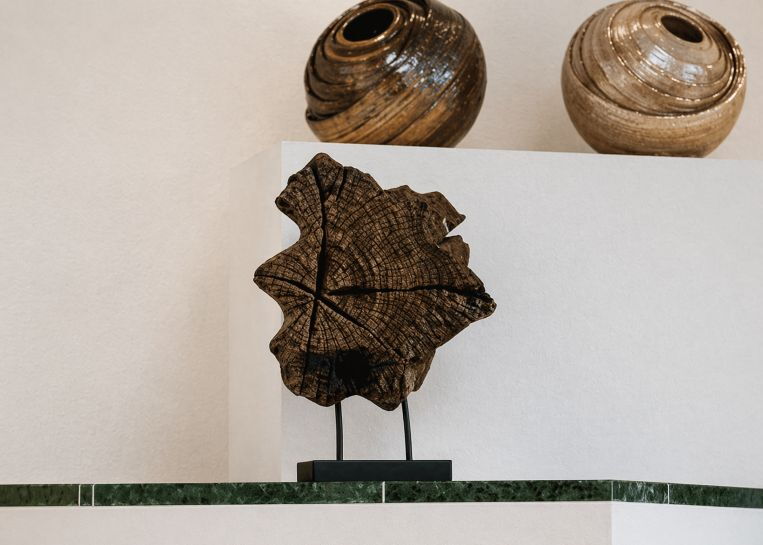 Detailaufnahme einer kunstvollen Altholz-Skulptur auf einem Metallsockel, platziert als Dekoration auf dem Vorsprung einer modernen Kaminanlage.