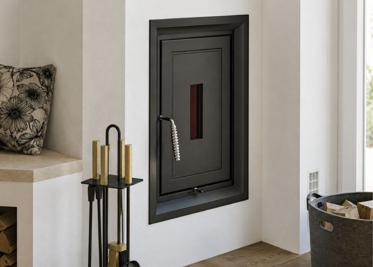 Nahaufnahme der schwarzen Gusstür mit kleinem Kontrollfenster und elegantem Griff, eingelassen in die weiße Ofenwand.