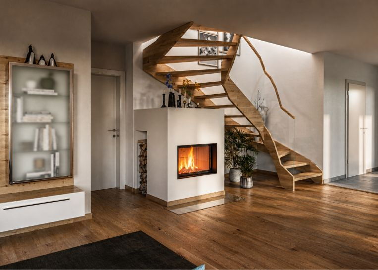 Ein kompakter, weiß verputzter Kamin mit Spartherm Varia ASh-4S-2 Einsatz, platziert unter einer modernen, geschwungenen Holztreppe im Wohnbereich.