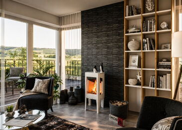 Gesamtansicht des weißen Jotul F163 Kaminofens vor einer dunklen Natursteinwand in einem hellen Wohnzimmer mit Balkonausblick.