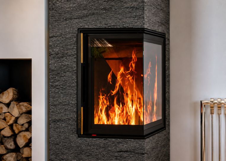 Nahaufnahme des brennenden Holzfeuers hinter dem großzügigen Eckglas, eingefasst in dunklen, strukturierten Naturstein.