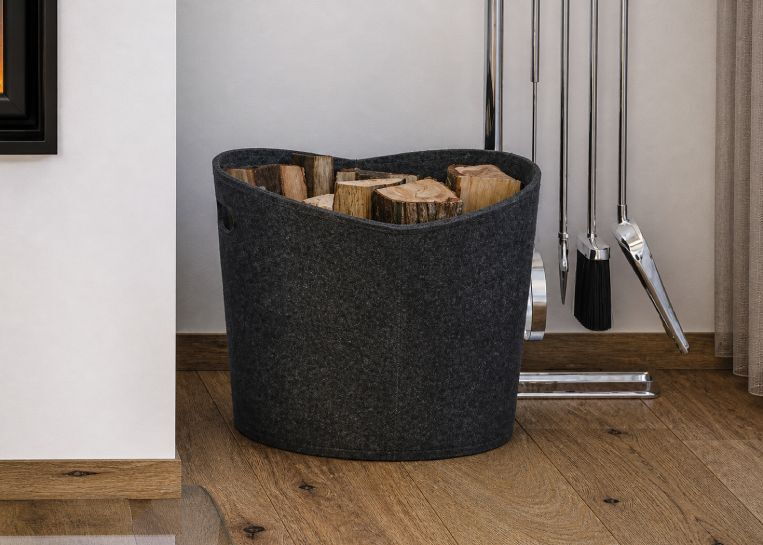 Ein anthrazitfarbener Filzkorb mit Brennholz neben dem schlichten Kaminbau und passendem Kaminbesteck.