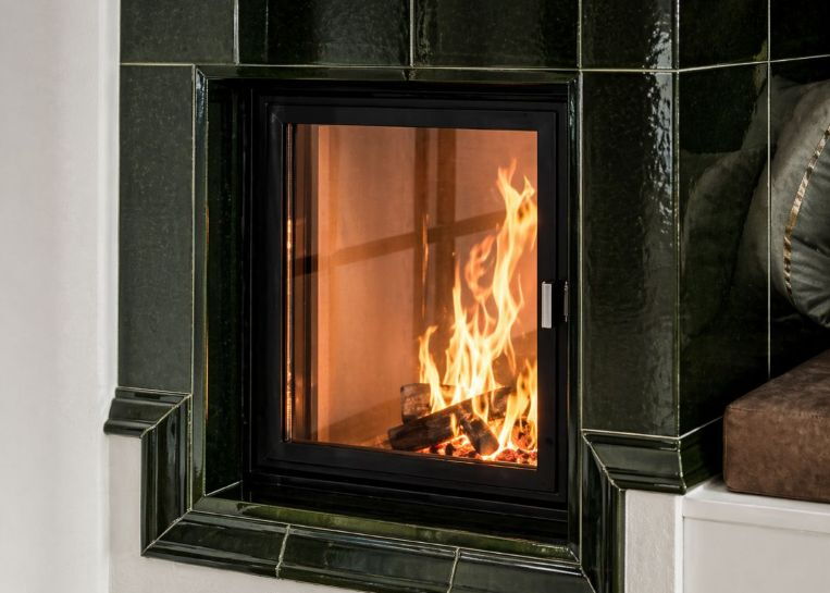 Nahaufnahme eines brennenden Holzfeuers hinter dem quadratischen Sichtfenster eines dunkelgrünen Kachelofens mit eckigen Keramikkacheln.