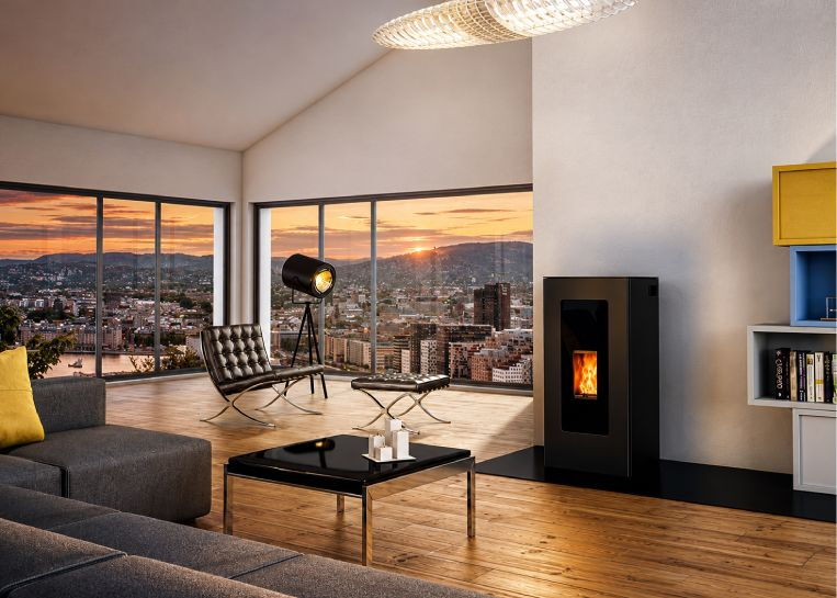 Ein schwarzer JØTUL PF 1230 Pelletofen in einem luxuriösen Penthouse-Wohnzimmer mit bodentiefen Fenstern und Stadtblick bei Sonnenuntergang.