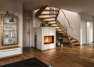 Ein kompakter, weiß verputzter Kamin mit Spartherm Varia ASh-4S-2 Einsatz, platziert unter einer modernen, geschwungenen Holztreppe im Wohnbereich.