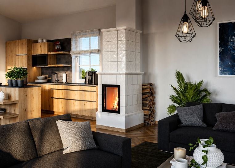 Ein heller Kachelofen mit dekorativem Aufsatz steht zentral zwischen einer modernen Holzküche und einem gemütlichen Wohnbereich mit dunklem Sofa.